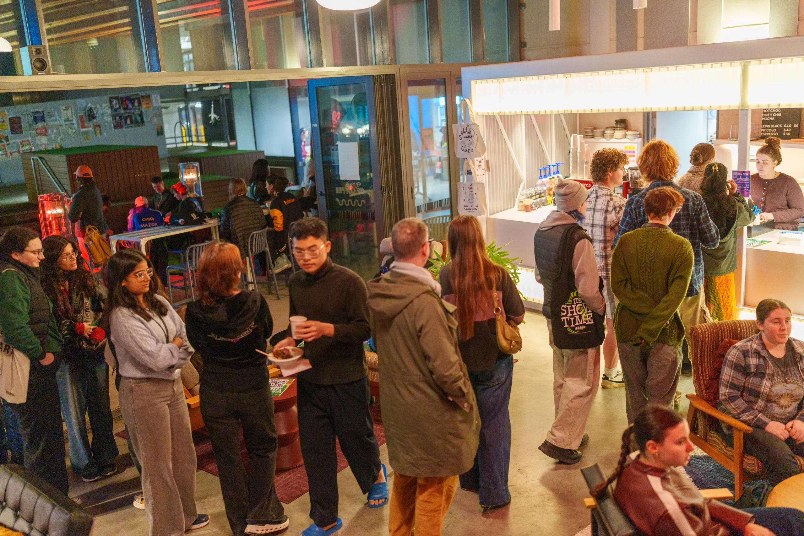 Students lining up for food at Grounded cafe as part of TUSA After Dark. In the background students sit around heaters and tables, and posters line the wall of a nearby fence.