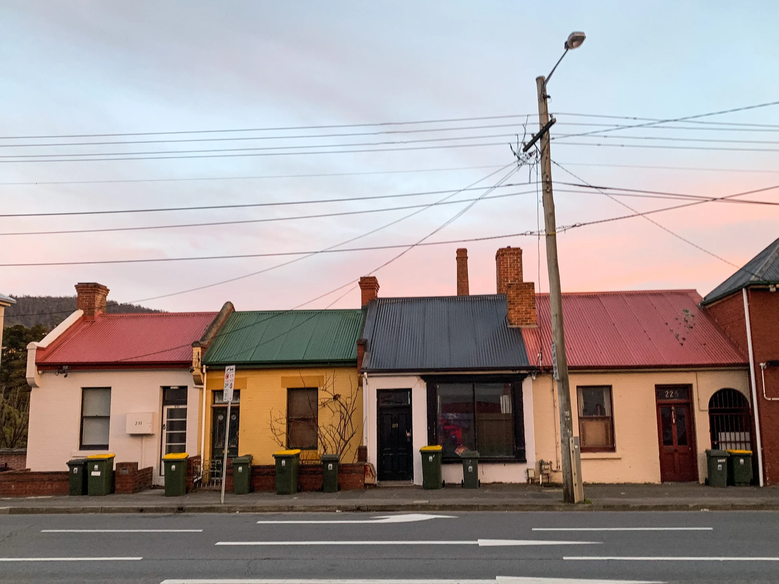A row of workers cottages sit next to each other, they are painted different colours: pink, yellow, white, with green and red rooves. Macquarie Street can be seen in the foreground, along with a telegraph pole.