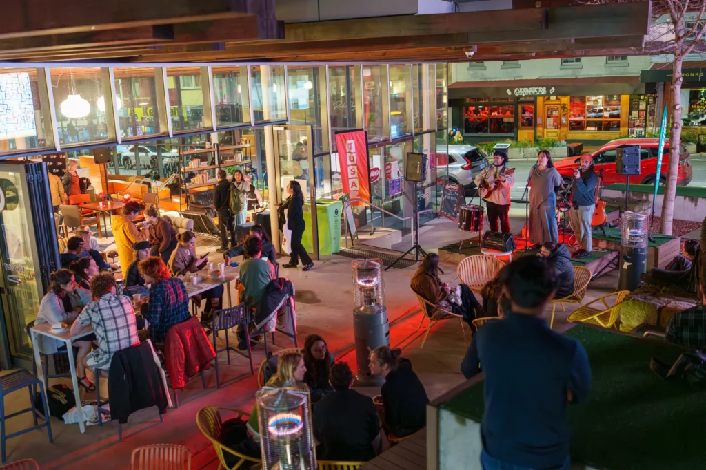 Musical trio Christian Palencia, Emily Oates and Alex Reid performing in the courtyard next to Grounded Cafe at the first TUSA After Dark. Students sitting at outdoor tables and around outdoor heaters watch on. Grinners bar can be seen across the road.