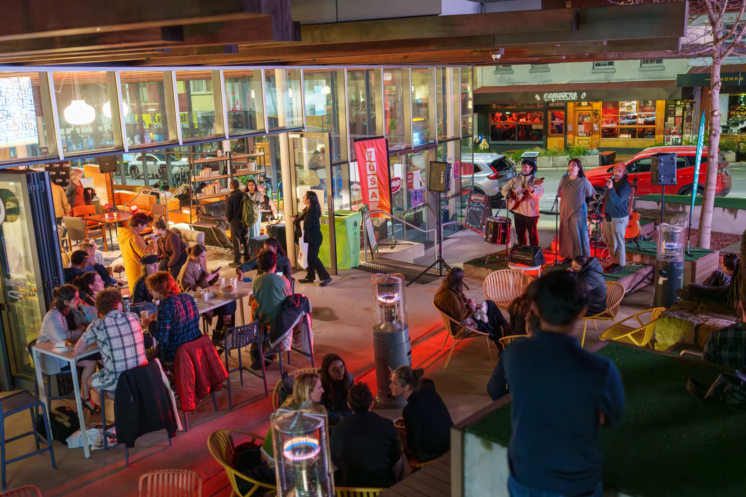 Musical trio Christian Palencia, Emily Oates and Alex Reid performing in the courtyard next to Grounded Cafe at the first TUSA After Dark. Students sitting at outdoor tables and around outdoor heaters watch on. Grinners bar can be seen across the road.