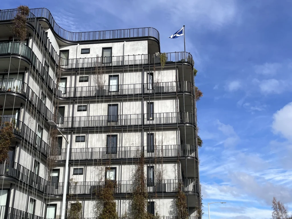 The Commons, a mid-rise housing option in the Hobart CBD. The building shown has curved balconies, plants growing on its exterior and a flag flying on the second top balcony.