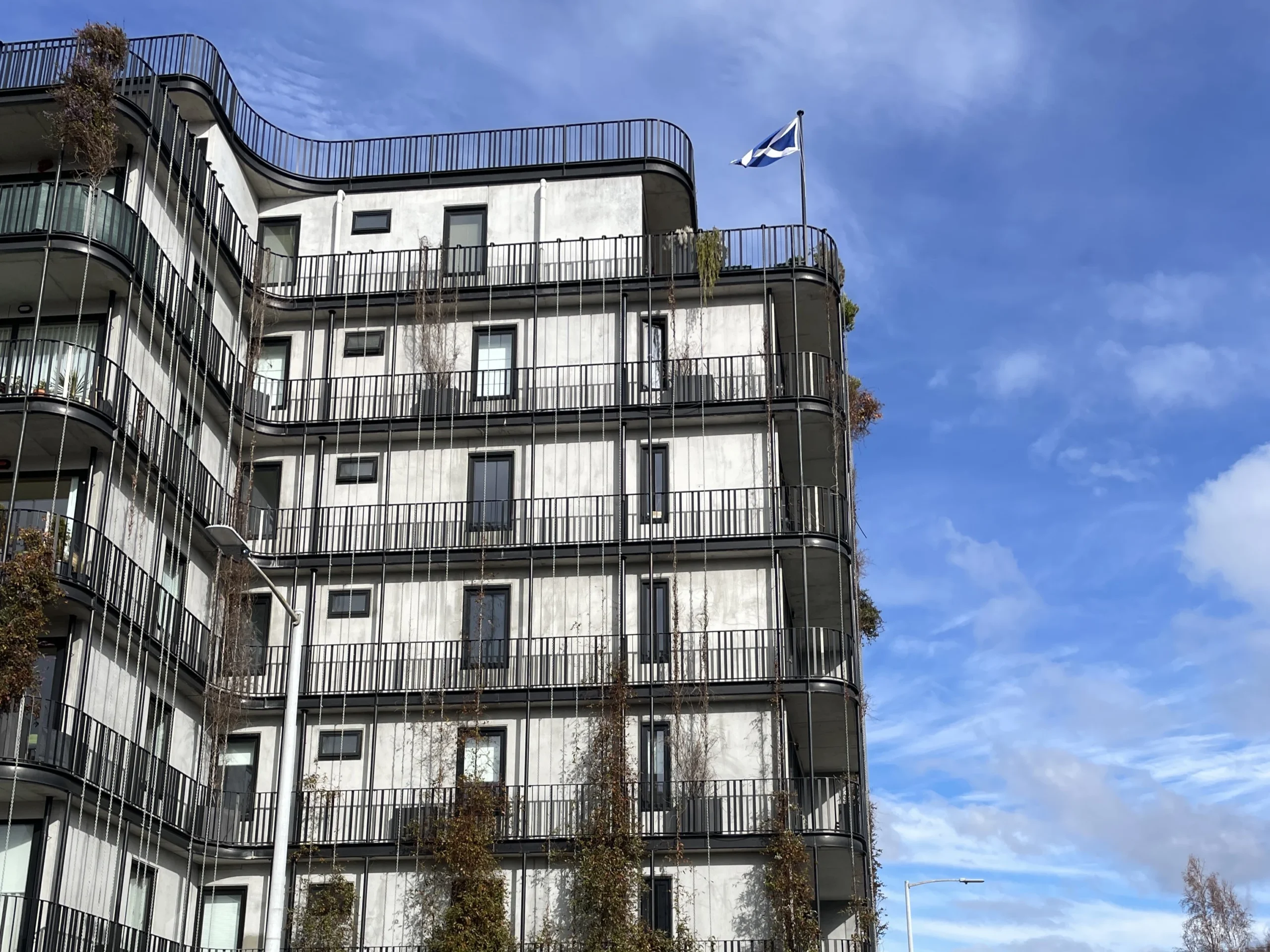 The Commons, a mid-rise housing option in the Hobart CBD. The building shown has curved balconies, plants growing on its exterior and a flag flying on the second top balcony.