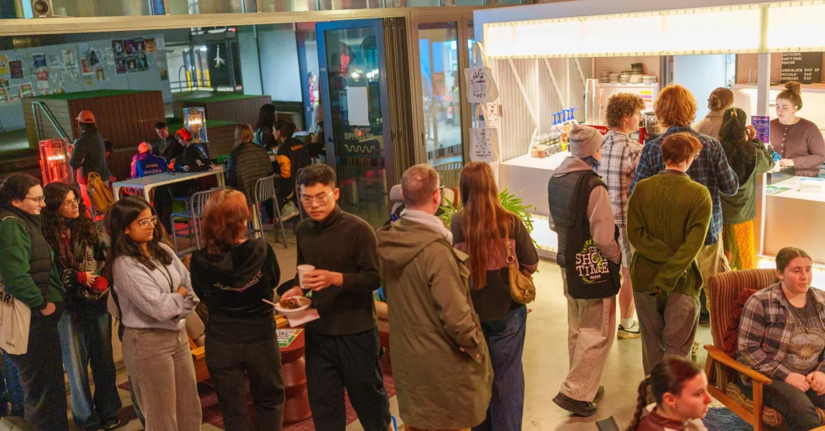 Students lining up for food at Grounded cafe as part of TUSA After Dark. In the background students sit around heaters and tables, and posters line the wall of a nearby fence.
