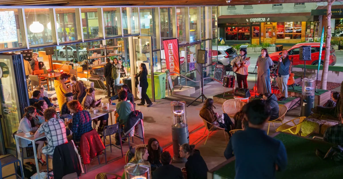 Musical trio Christian Palencia, Emily Oates and Alex Reid performing in the courtyard next to Grounded Cafe at the first TUSA After Dark. Students sitting at outdoor tables and around outdoor heaters watch on. Grinners bar can be seen across the road.