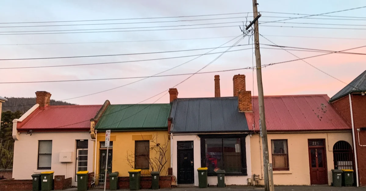 A row of workers cottages sit next to each other, they are painted different colours: pink, yellow, white, with green and red rooves. Macquarie Street can be seen in the foreground, along with a telegraph pole.